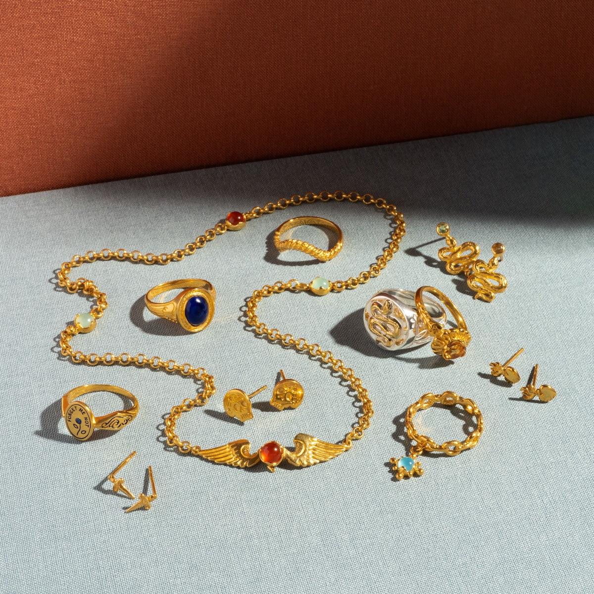 A selection of gold and silver jewelry, including 18k yellow gold vermeil rings, necklaces, cufflinks, and the Serpent Stud Earrings, displayed on a light blue surface with an orange background. Some feature tourmaline gemstones and intricate designs.