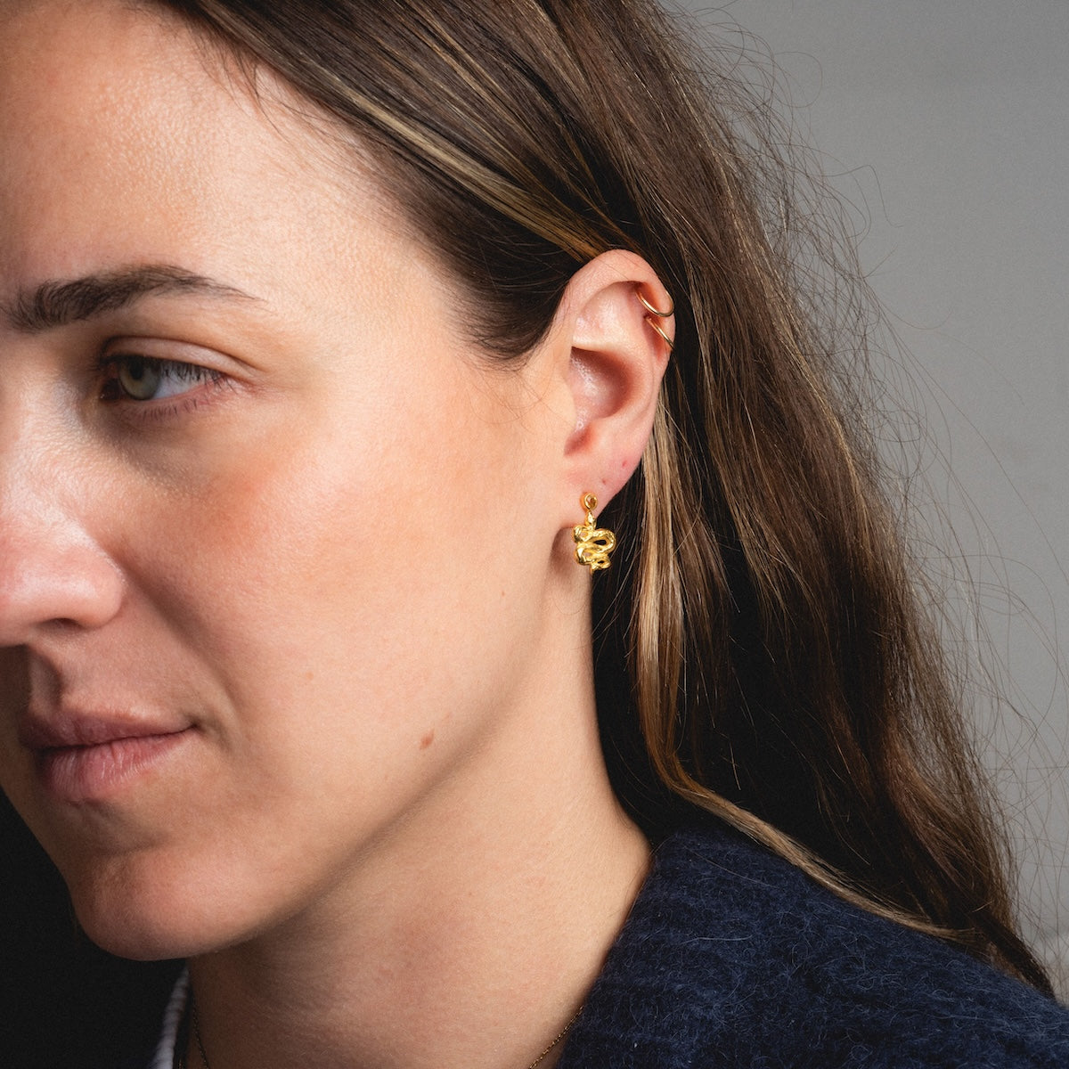 A close-up shows a woman with long brown hair and light skin wearing gold earrings and Serpent Stud Earrings. She has a neutral expression, wears a dark blue top, and stands against a plain gray background.