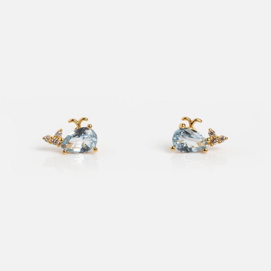 The Whale Crystal Studs by Tai Jewelry are gold earrings shaped like whales, featuring light blue gemstones as bodies and sparkling tail accents, displayed on a plain white background.