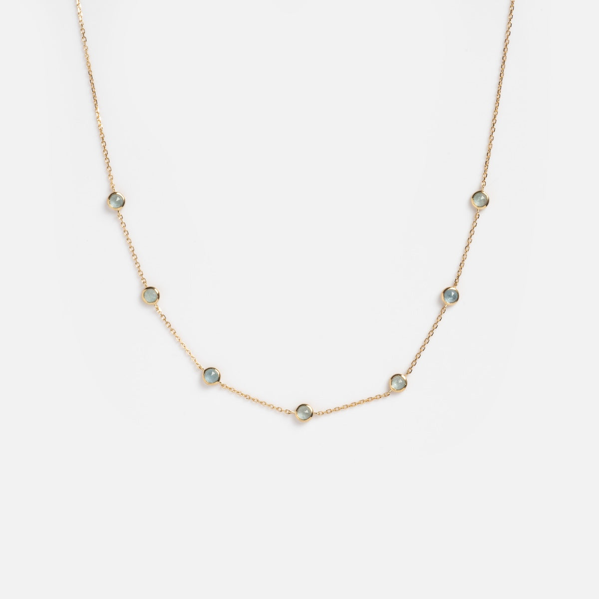 The Solid Gold Birthstone Station Necklace Sample is a 9k yellow gold chain with seven bezel-set, round lab-created blue gemstones, elegantly spaced along the length and displayed on a plain white background.