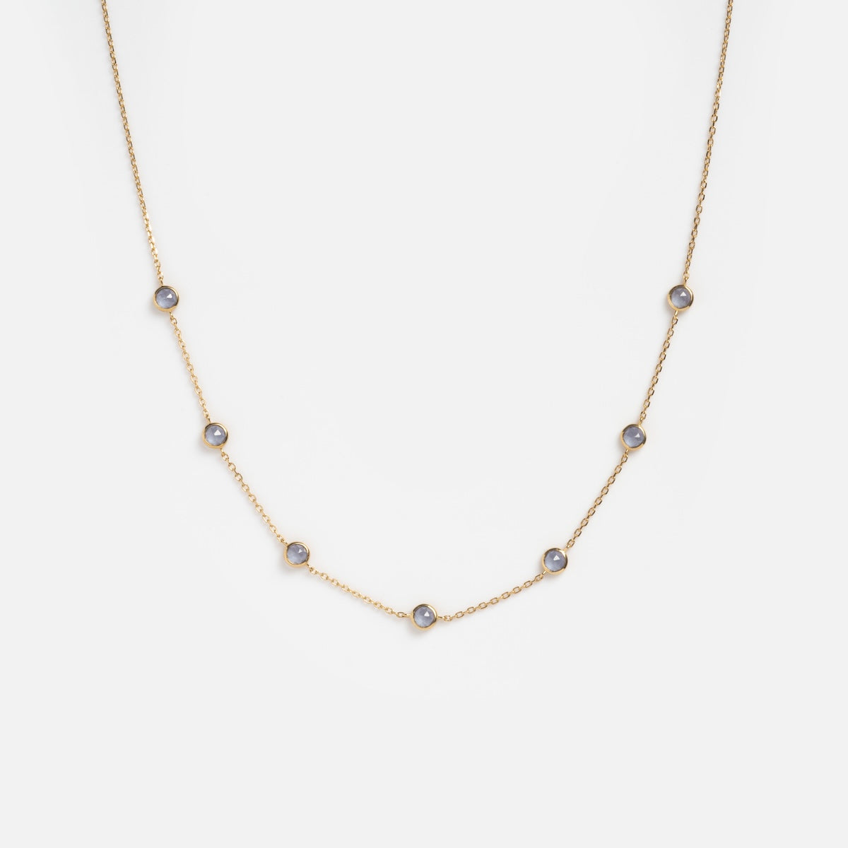 The Solid Gold Birthstone Station Necklace Sample is a delicate 9k yellow gold necklace with seven evenly spaced round lab-created gemstones in bezel settings, shown on a plain white background.