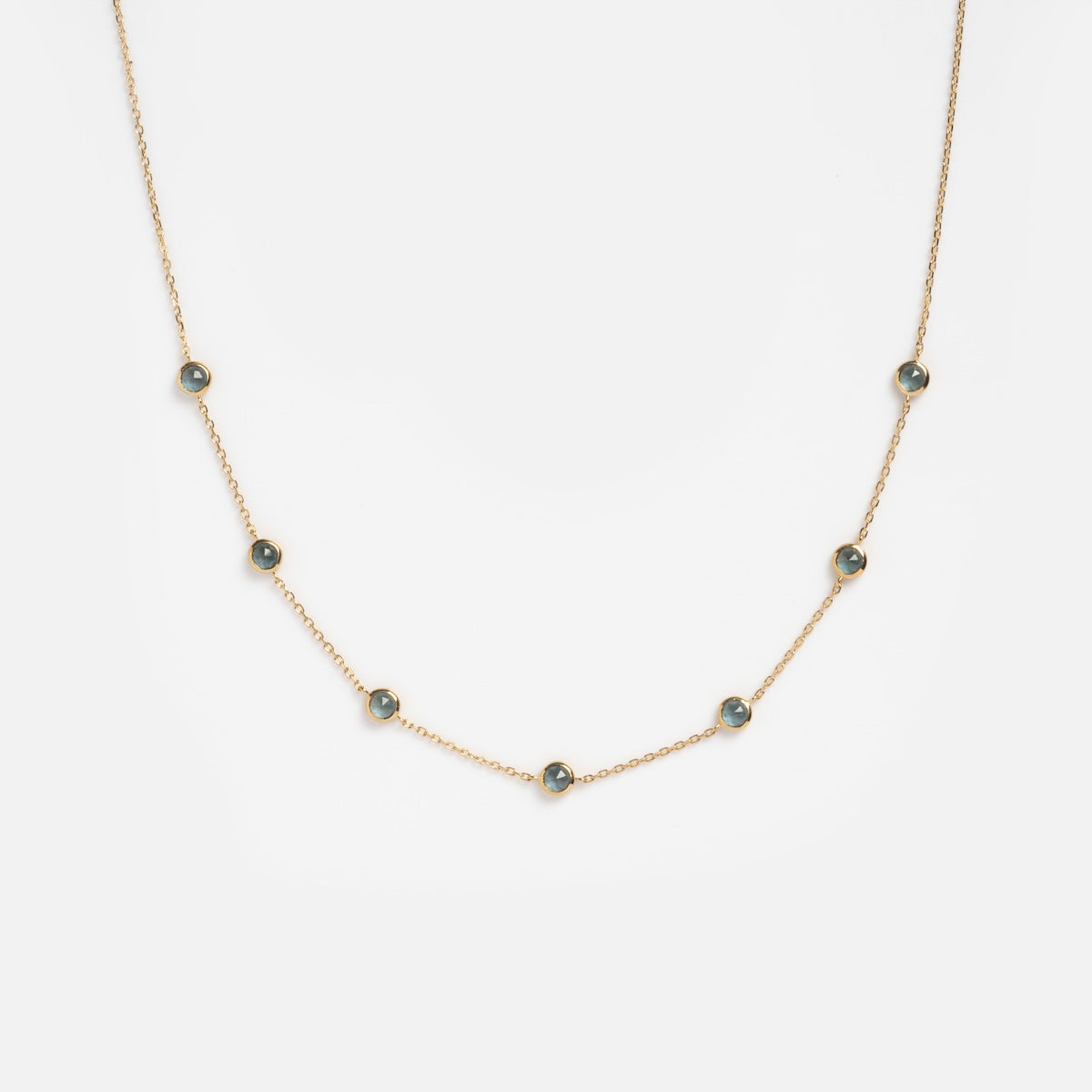 The Solid Gold Birthstone Station Necklace Sample features seven small blue lab-created gemstones in round bezels, evenly spaced on a 9k solid yellow gold chain. The simple, elegant design stands out against a plain white background.