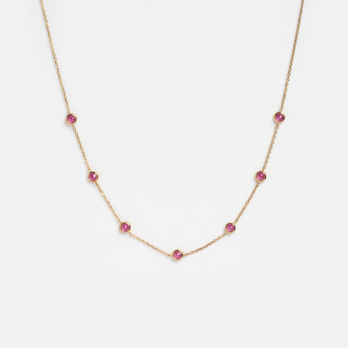 The Solid Gold Birthstone Station Necklace Sample is a delicate 9k yellow gold chain with seven evenly spaced lab-created pink gemstones, shown on a plain white background.