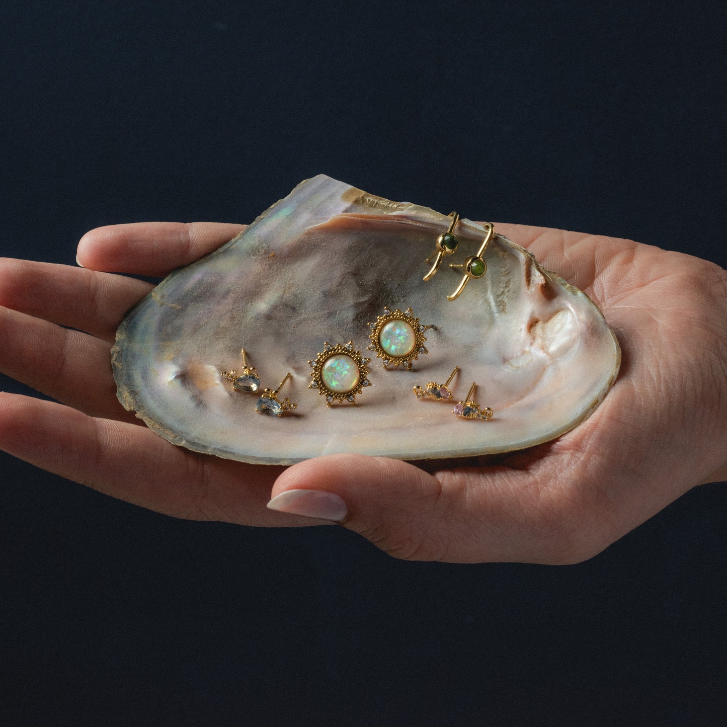 A hand displays an iridescent oyster shell holding three pairs of 14k gold plated earrings, including the Dillon Studs, each featuring opal-like stones and intricate settings, set against a dark background.