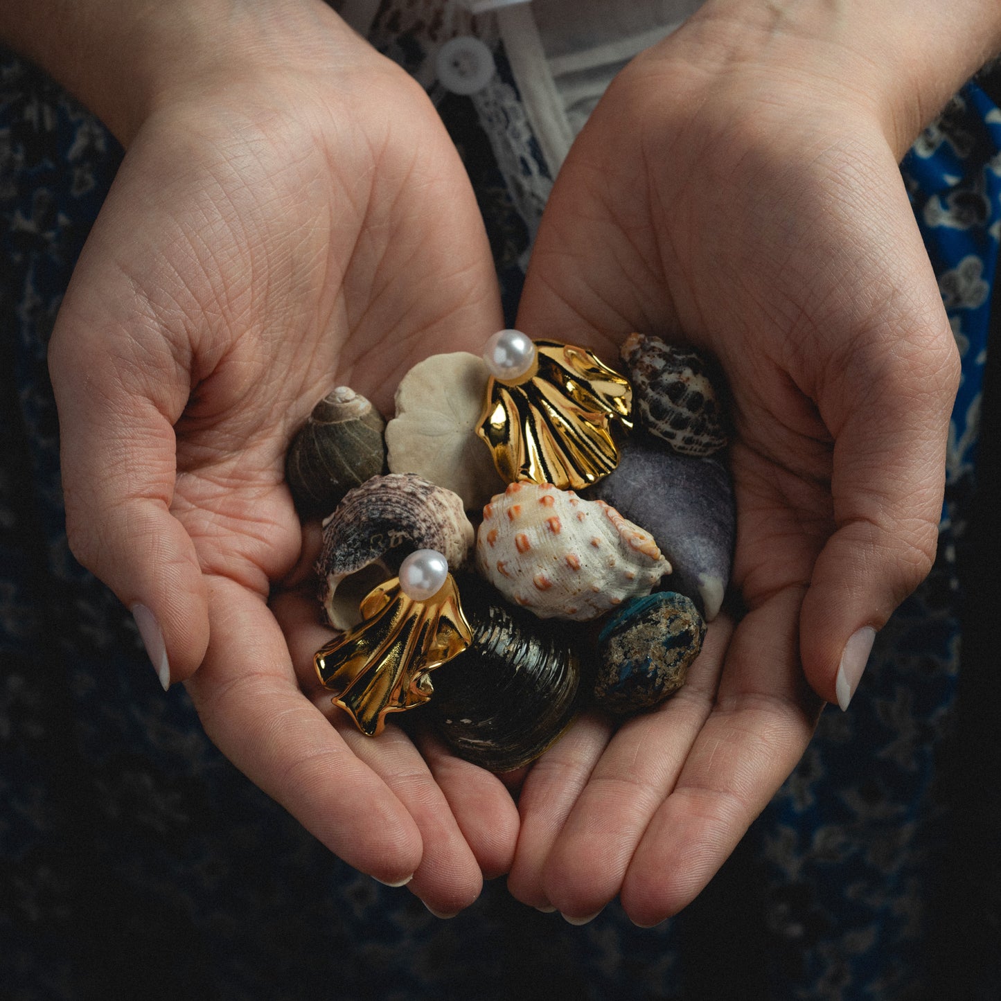 A pair of hands gently cupping various seashells and two versatile Waves Earrings—gold shell-shaped glass pearl earrings—against a blurred background.