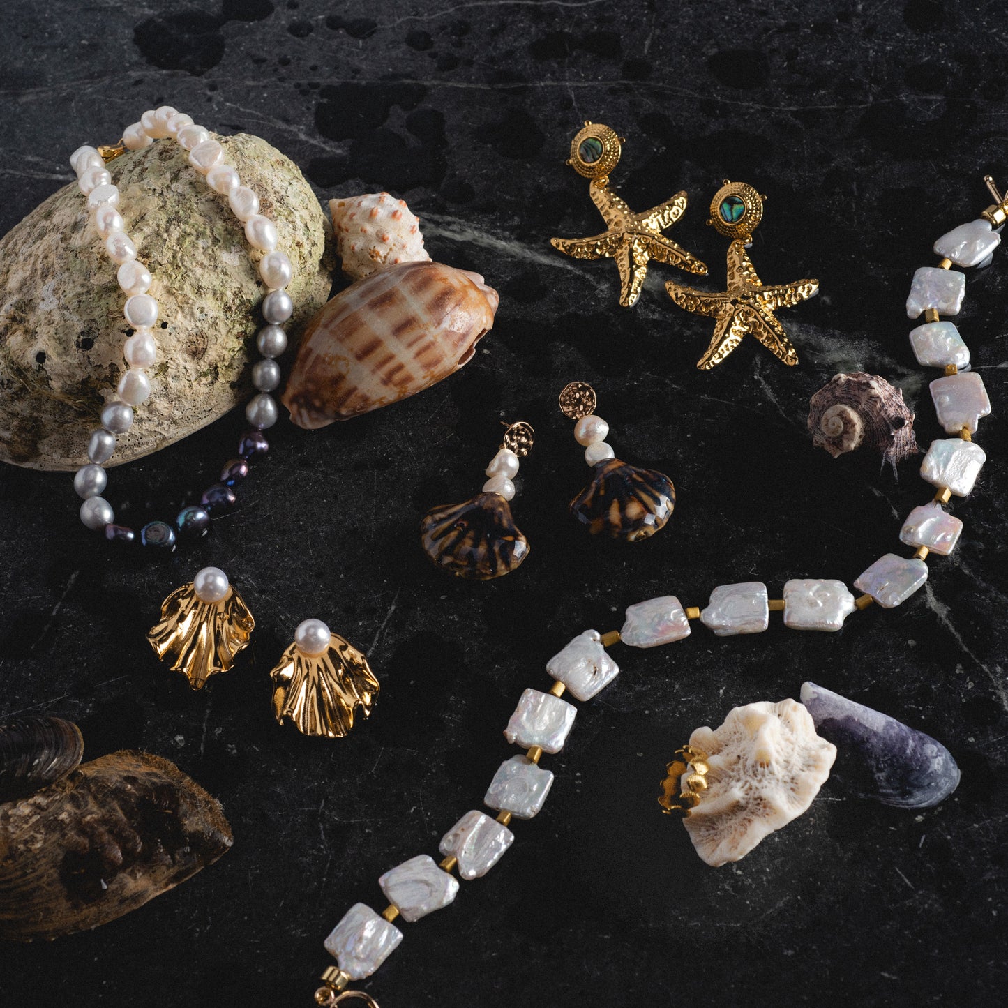 A Seashells Ring and other jewelry items, such as pearl necklaces, gold starfish and shell-shaped earrings, plus a pearl bracelet, are displayed on a dark stone surface.