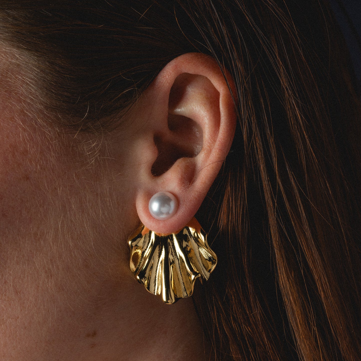 Close-up of a person with straight, reddish-brown hair wearing Waves Earrings—elegant pearl studs with round white glass pearls and large gold shell-shaped dangles. These versatile earrings add charm to any look.
