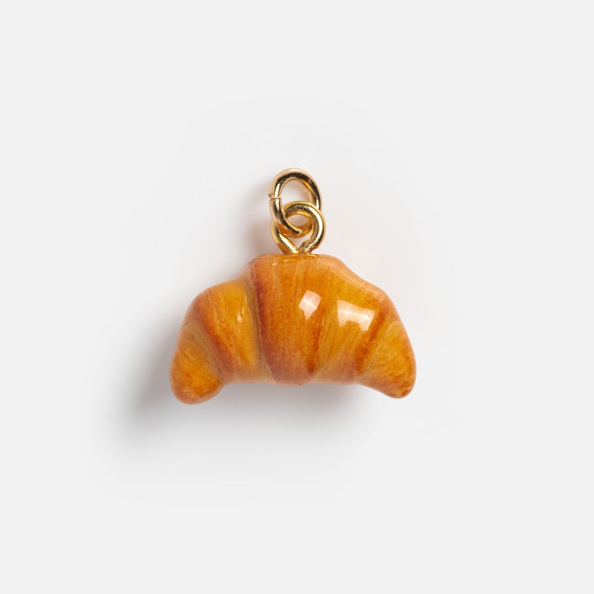 The Charms by Warm Clay Co features a small, shiny croissant-shaped charm, hand-crafted from gold plated clay with a golden loop for attaching to bracelets or necklaces, displayed against a plain white background.