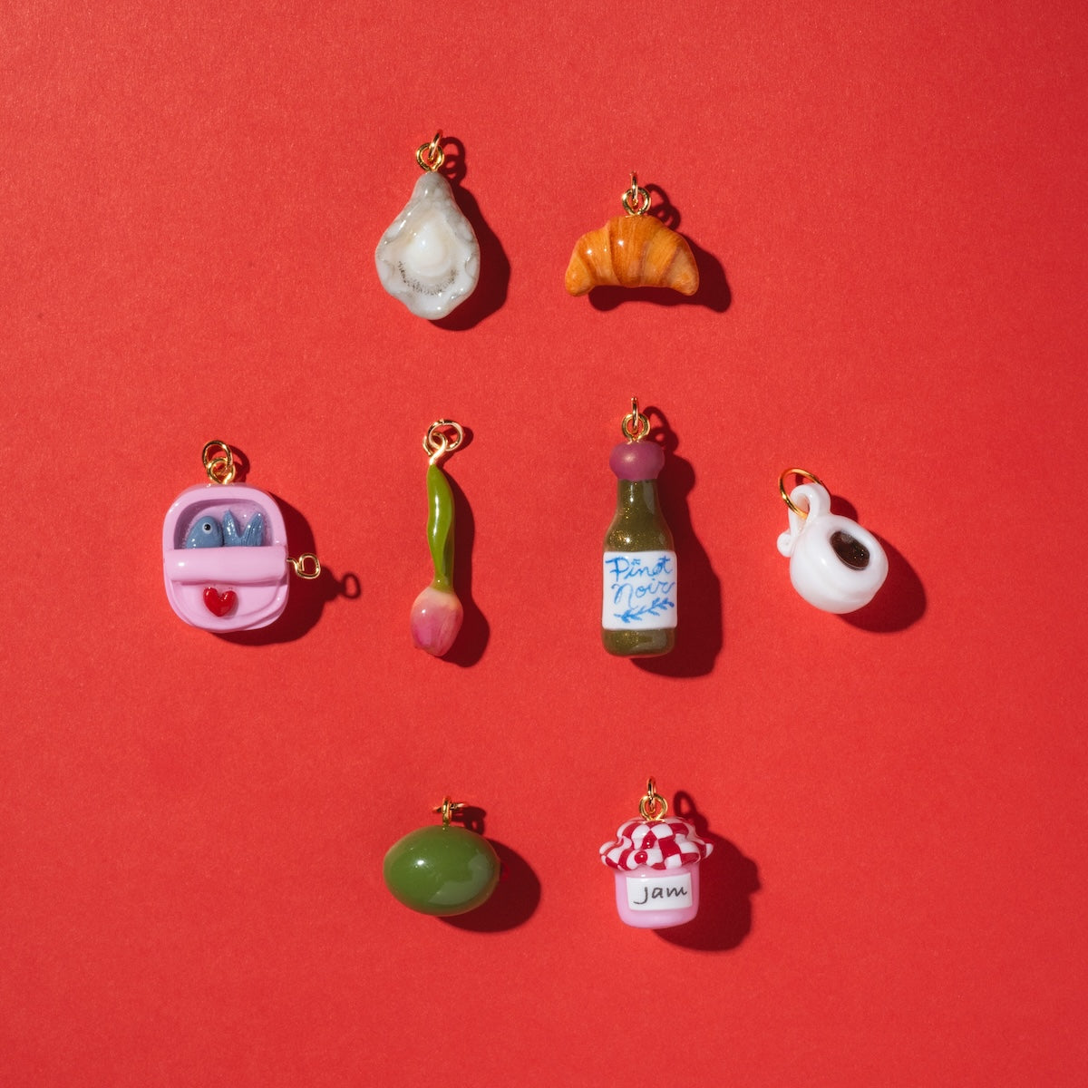 Charms by Warm Clay Co features nine hand-crafted charms on a red background, including an oyster, croissant, toaster, green spoon, wine bottle, swan, green olive, and a jar labeled jam—perfect unique necklace charms.