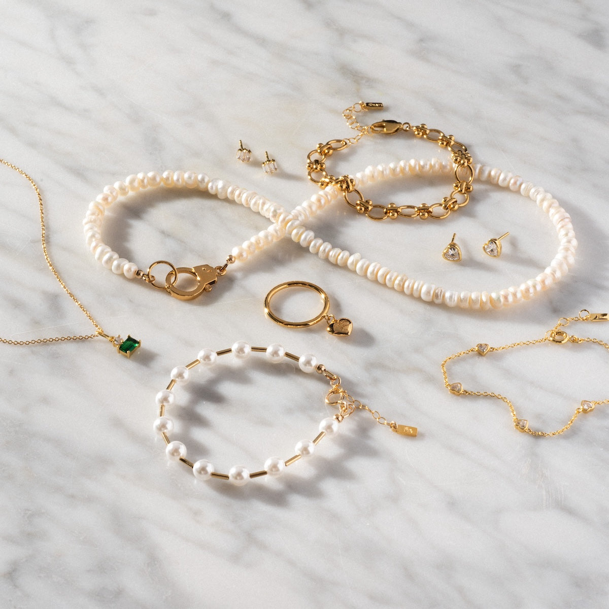 The All Of My Hearts Bracelet—an 18k yellow gold plated simulated diamond piece—is displayed alongside assorted gold and pearl necklaces, earrings, and rings on a white marble surface with soft lighting.