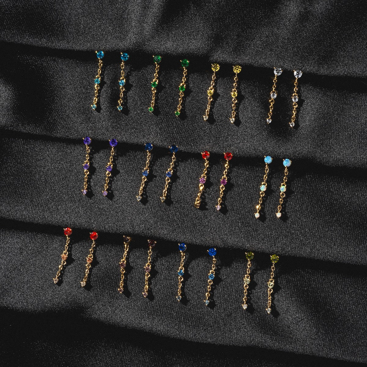 The Celebration Ombre Birthstone Drop Studs are vibrant 14k gold plated earrings featuring round, multicolored gemstones, elegantly arranged in neat rows on a textured black fabric background.