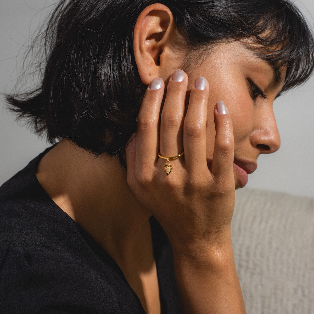 A woman with short dark hair, wearing a black top, rests her chin on her hand and showcases the Sweet Heart Ring—an elegant piece featuring an 18k gold-plated band with a delicate heart-shaped charm. She sits on a light textured surface.