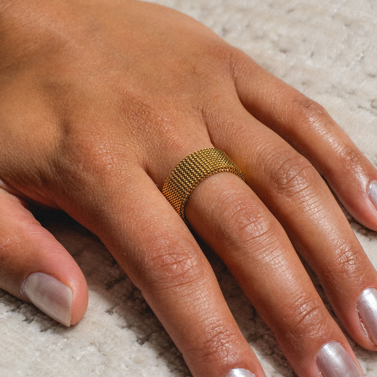 A hand with light brown skin wears the Priscila Ring, an 18k yellow gold plated statement piece with a textured design, on the middle finger. The hand rests on a light, textured surface and the nails have a metallic light polish.