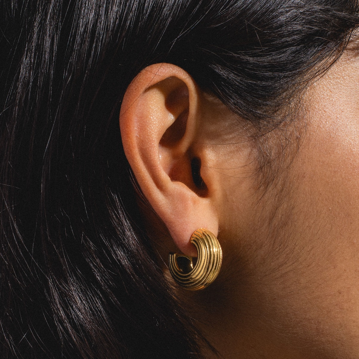 Close-up of a medium-toned person wearing Jerry Earrings—chunky gold earrings with a layered, textured style. Dark, straight hair partially covers the ear.