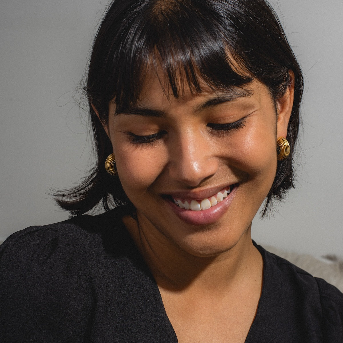 A woman with short dark hair, wearing Jerry Earrings and a black top, smiles gently while looking down against a plain background.