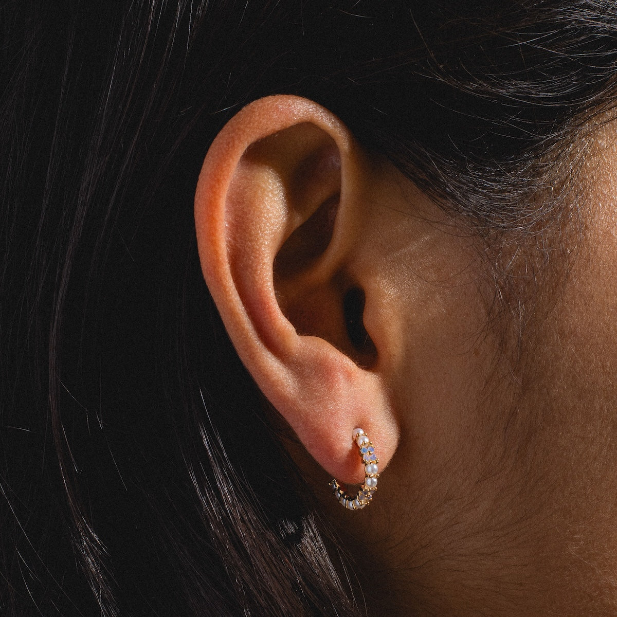 A close-up of a person with medium to dark skin and dark brown hair wearing Orchid Pearl Hoops—small, gold-plated hoops featuring sparkling stones.