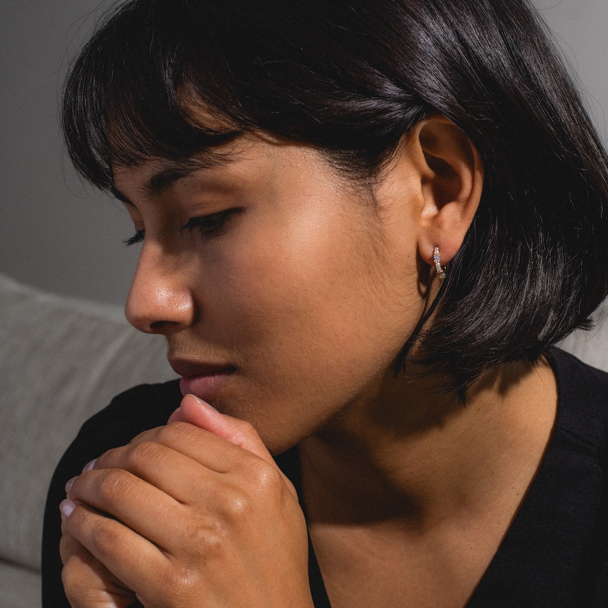 A woman with short dark hair wears Orchid Pearl Hoops as she sits indoors on a light-colored couch, eyes closed and hands clasped near her face, wearing a black top and appearing deep in thought.