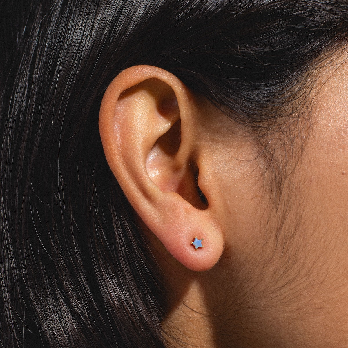 Close-up of a person’s ear with straight dark hair, wearing Milky Blue Star Studs—small celestial earrings shaped like stars.