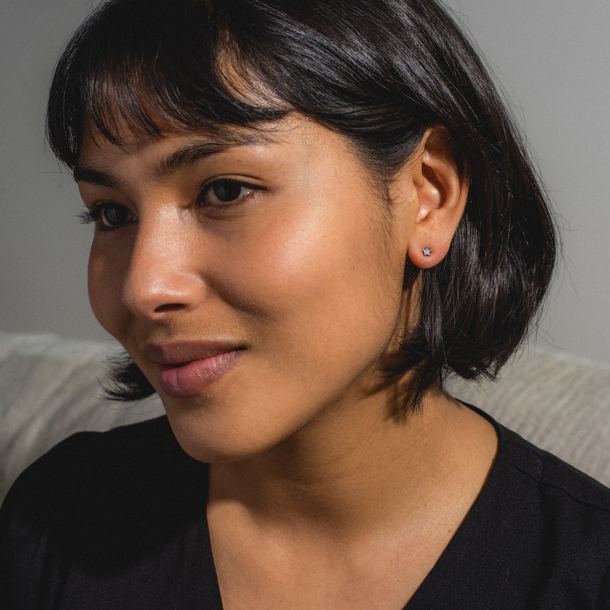 A woman with short dark hair and side-swept bangs smiles softly, wearing a black top and Milky Blue Star Studs, sitting on a light-colored sofa with a neutral background.