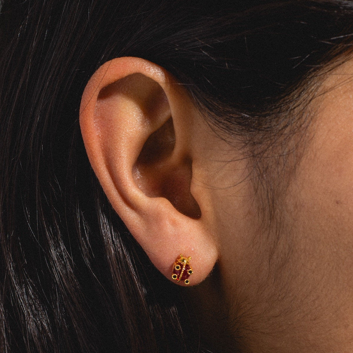 Close-up of a persons ear with straight dark hair, wearing Ladybug Studs in red and gold.