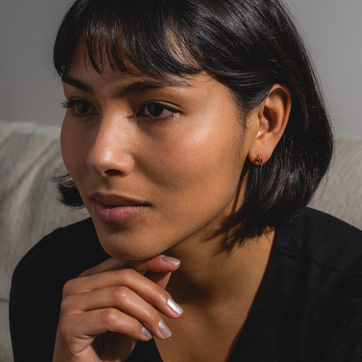 A woman with short, dark hair and side-swept bangs sits indoors, resting her chin on her hand. She wears a black top and the Ladybug Studs, looking thoughtfully to the side.