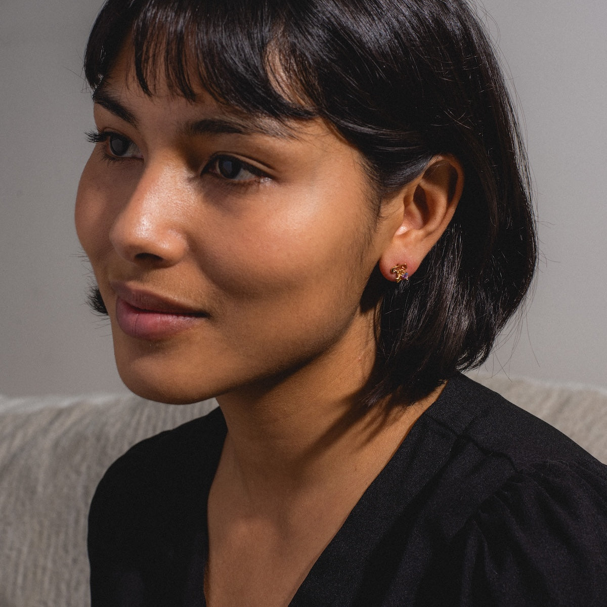 A woman with short dark hair and bangs, wearing a black top and the Pillow Talk Dangle Earrings—18k gold plated earrings shaped like small flowers—sits on a light-colored couch, gazing gently to the side.