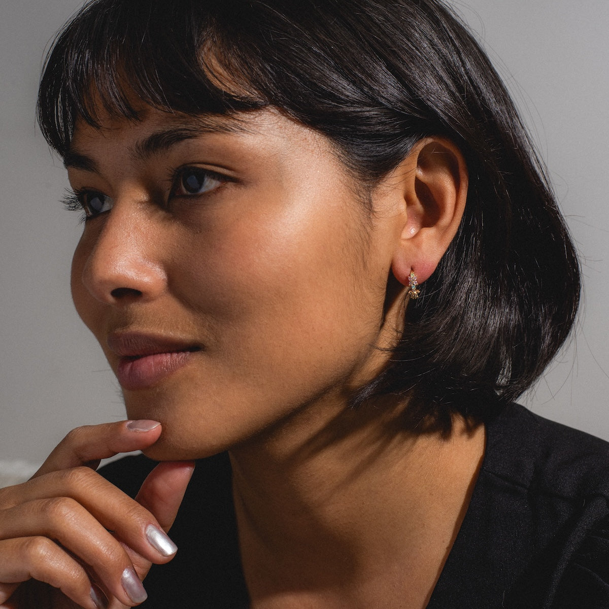 A woman with short dark hair and glowing skin gazes thoughtfully to the side, resting her chin on her hand. She wears a black top, subtle makeup, and Pastel Star Huggie Hoops that add a touch of sparkle to her look.
