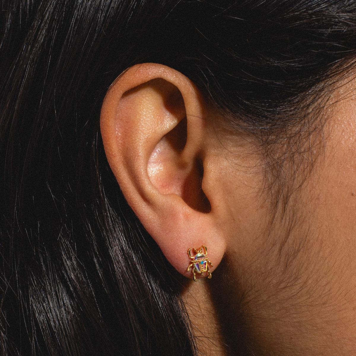 Close-up of a person’s ear wearing Beetlejuice Studs—18k gold plated beetle-shaped earrings with iridescent CZ stones. Part of the persons dark hair is visible.