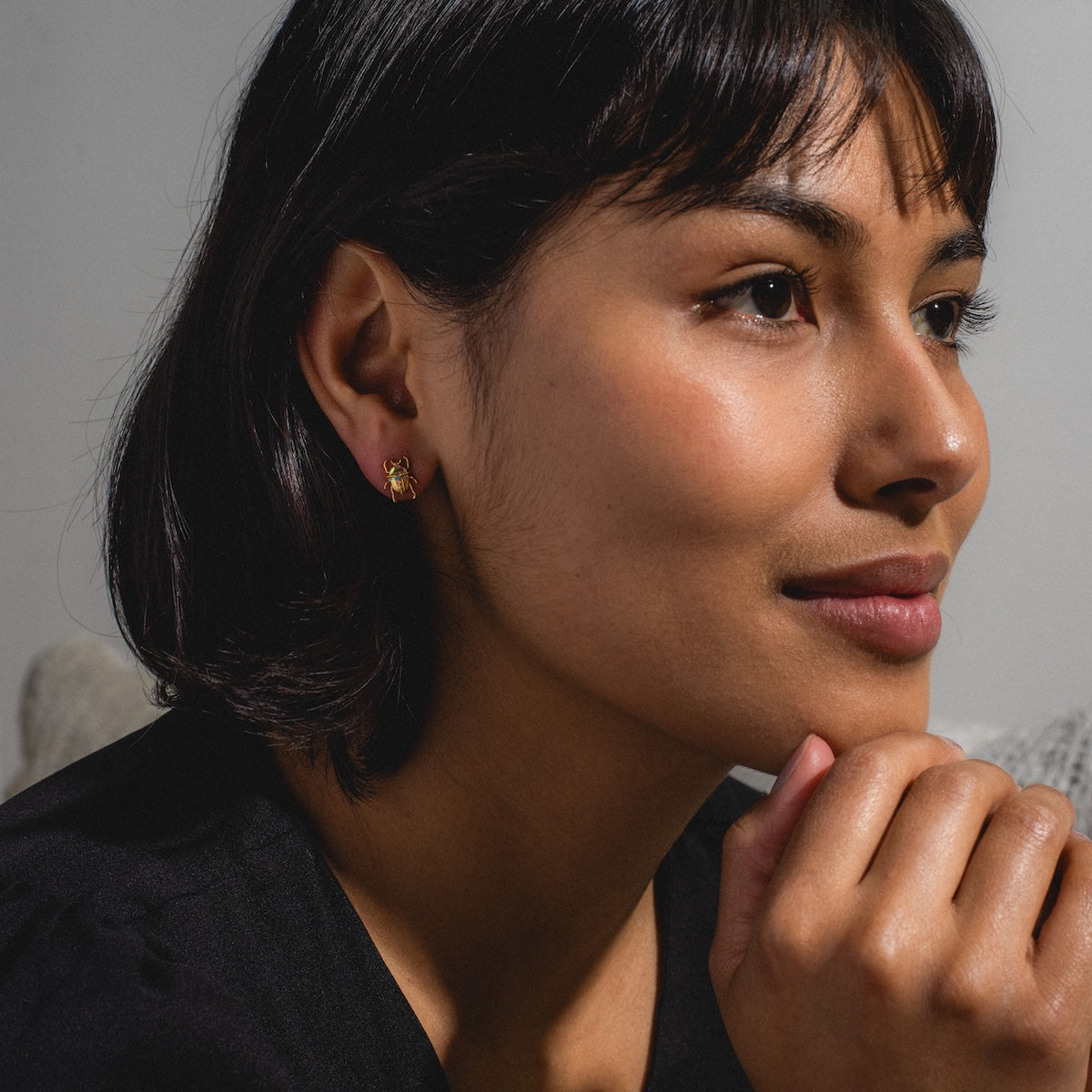 A woman with short dark hair wears Beetlejuice Studs and a black top, resting her chin on her hand and gazing thoughtfully to the side in a softly lit, calm setting.