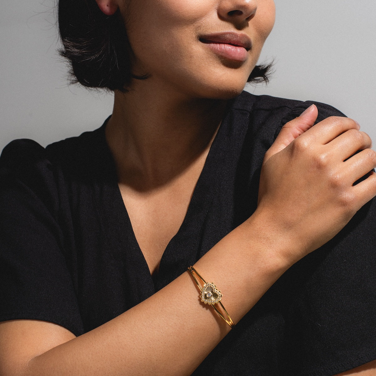 A woman with short dark hair, wearing a black top and the "In the Name of Love Cuff Sample"—an 18k yellow gold plated bracelet with a charm—crosses her arm over her chest and smiles softly against a neutral background.