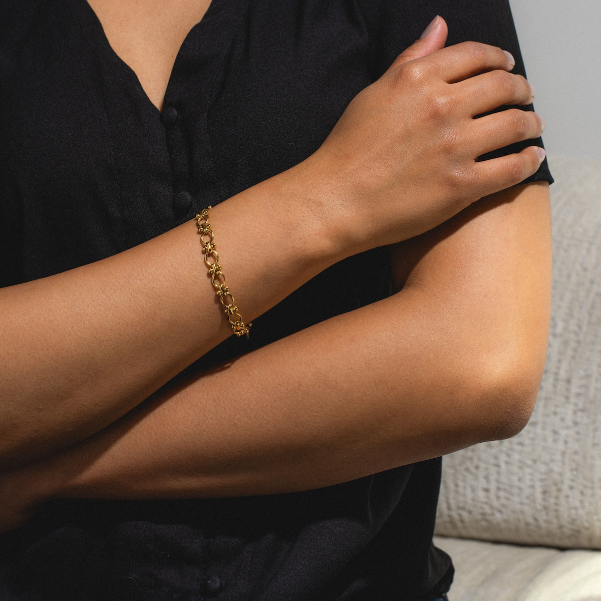 A person wearing a black short-sleeve shirt sits on a light-colored couch with their arms crossed, showcasing the Aubrey Bracelet on their wrist.