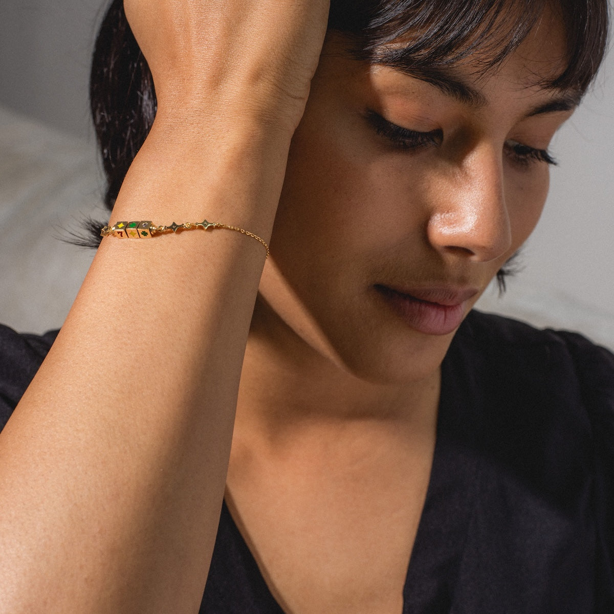 A person with short, dark hair and a black top rests their head on their hand, displaying the Hit The Jackpot Bracelet in 18k yellow gold plating with delicate green and yellow stones on their wrist.