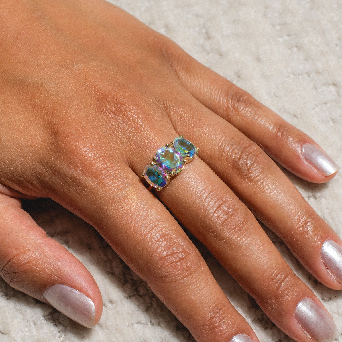 A hand with light brown skin wears the One of a Kind 14k Angel Aura Gumdrop Ring, set with three iridescent gemstones. Resting on a textured surface, metallic silver nails highlight this unique jewelry piece.