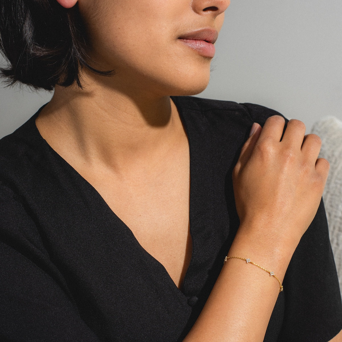 A woman in a black top touches her shoulder, revealing the Shimmer Blossom Bracelet, an elegant 18k gold plated accessory, on her wrist. Her face is partly visible as she poses against a light background.
