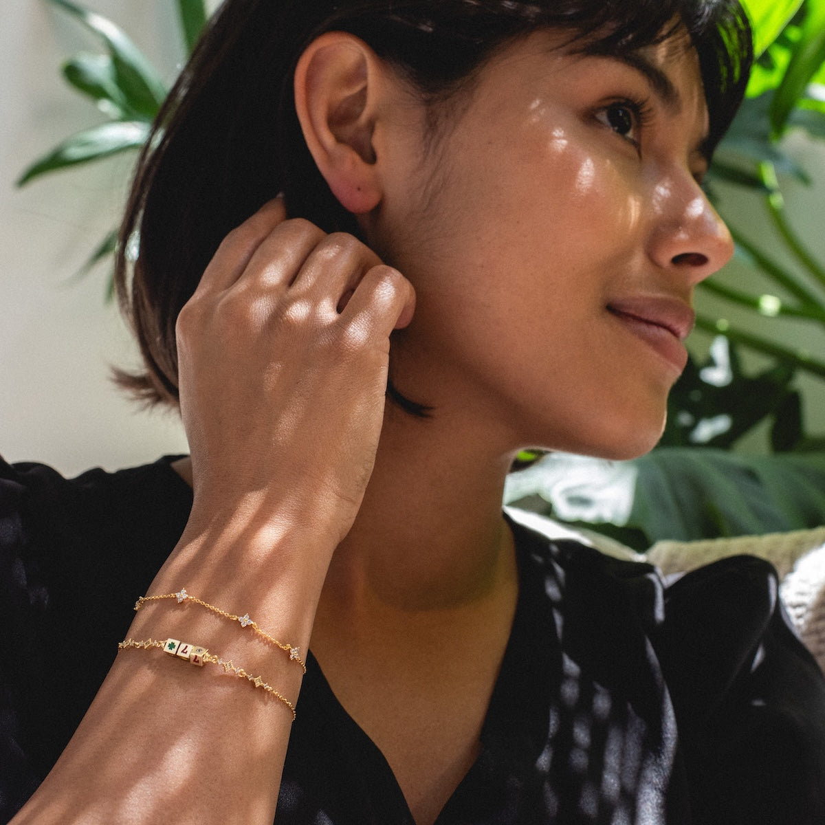 A woman with short dark hair sits in sunlight, wearing a black top and two delicate Hit The Jackpot Bracelets. She touches her ear as dappled light and plant leaves create a dreamy background.