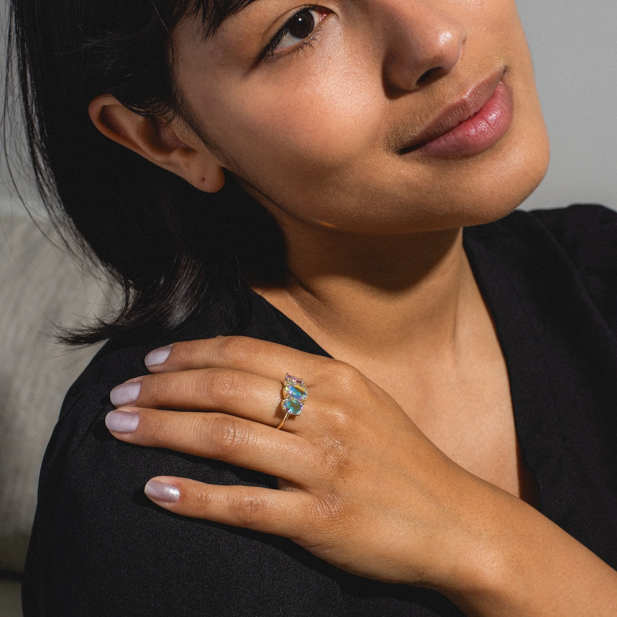 A woman with short dark hair and a soft smile rests her hand on her shoulder, showing off the One of a Kind 14k Angel Aura Gumdrop Ring, set with colorful gemstones. She wears a black top and natural makeup.