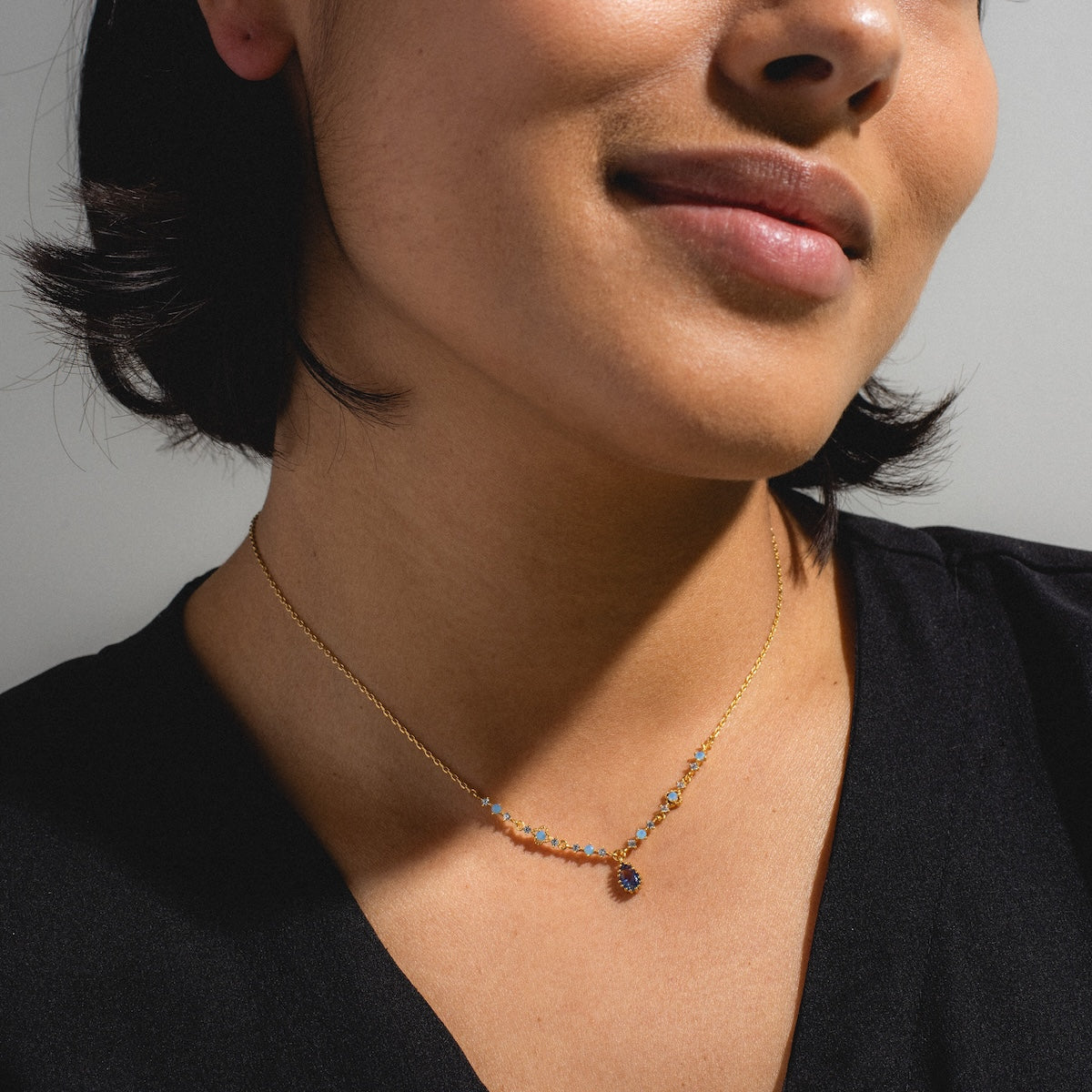 A woman wearing a black top and the Something Blue Necklace Sample, a delicate 18k yellow gold plated brass necklace with colorful gemstones, poses against a neutral background.