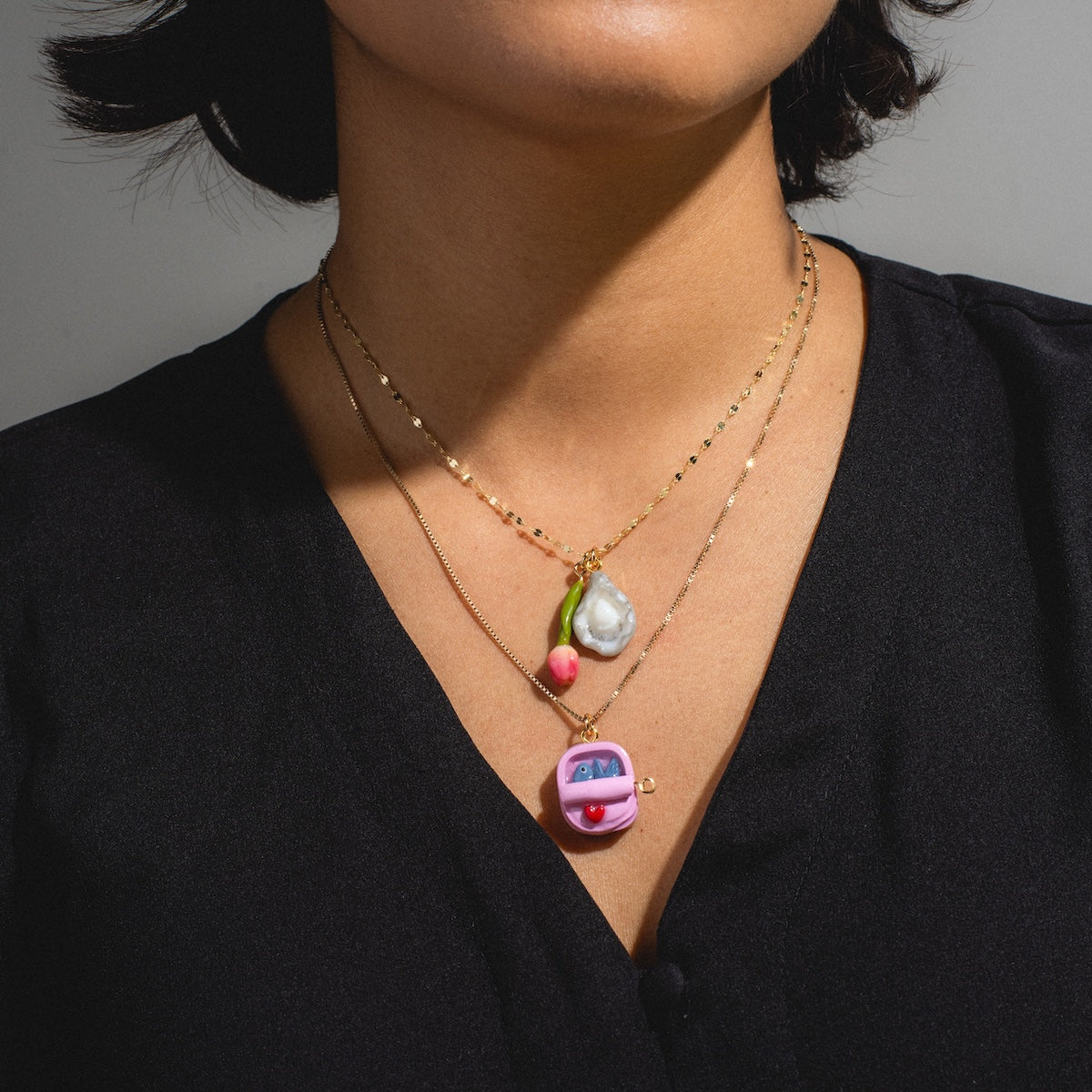 A person wears a black V-neck top and three layered gold necklaces from Charms by Warm Clay Co, each featuring hand-crafted charms: a pink van, a pearl-like shape, and a small pink bead. The head is partially out of frame.