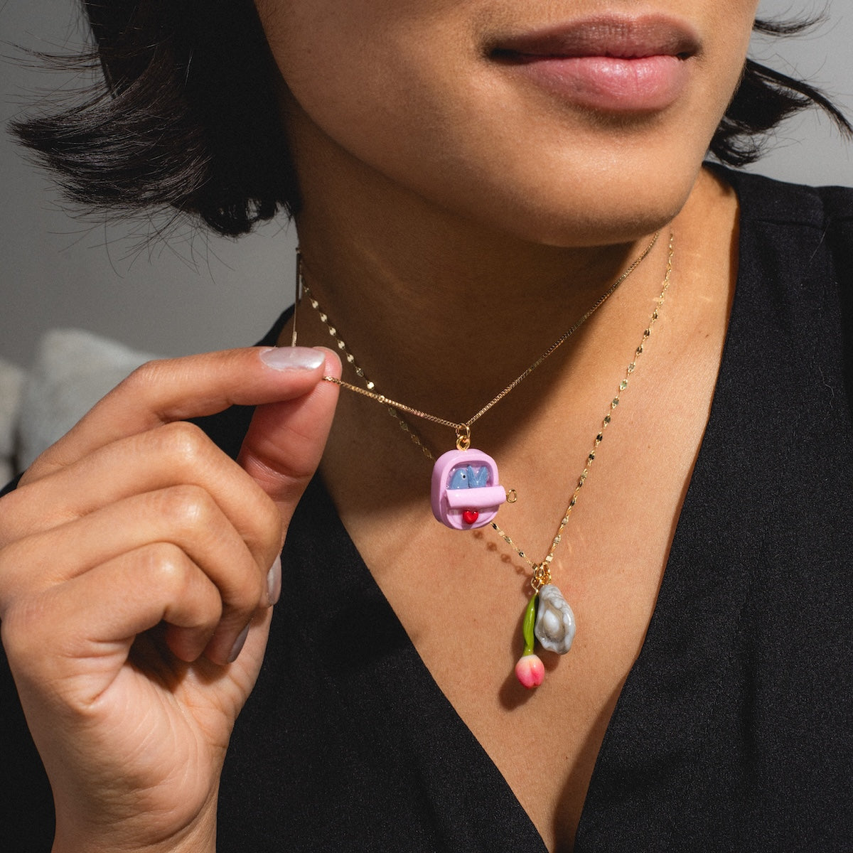 A woman in a black top holds up a gold Charms by Warm Clay Co necklace with a pink bus pendant and unique charms, and also wears another gold necklace with oyster and cherry-shaped pendants.