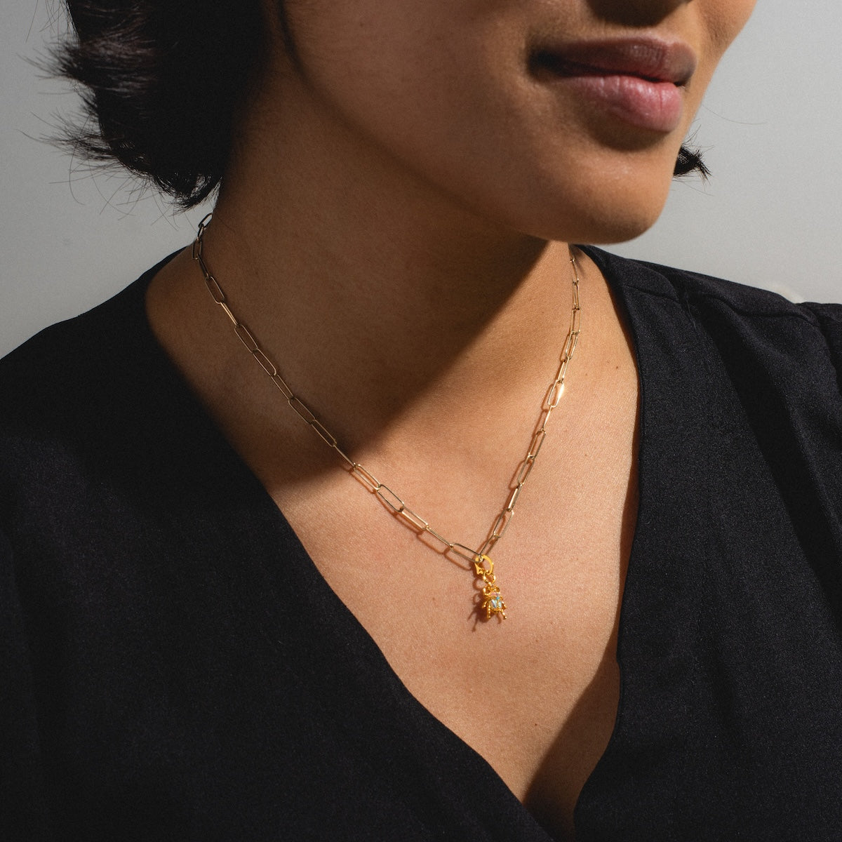 A woman in a black V-neck top models the Charms by Girls Crew necklace, featuring a gold chain with elongated links and an 18k yellow gold Beetlejuice Charm pendant. The cropped image highlights her neck, collarbone, and part of her face.