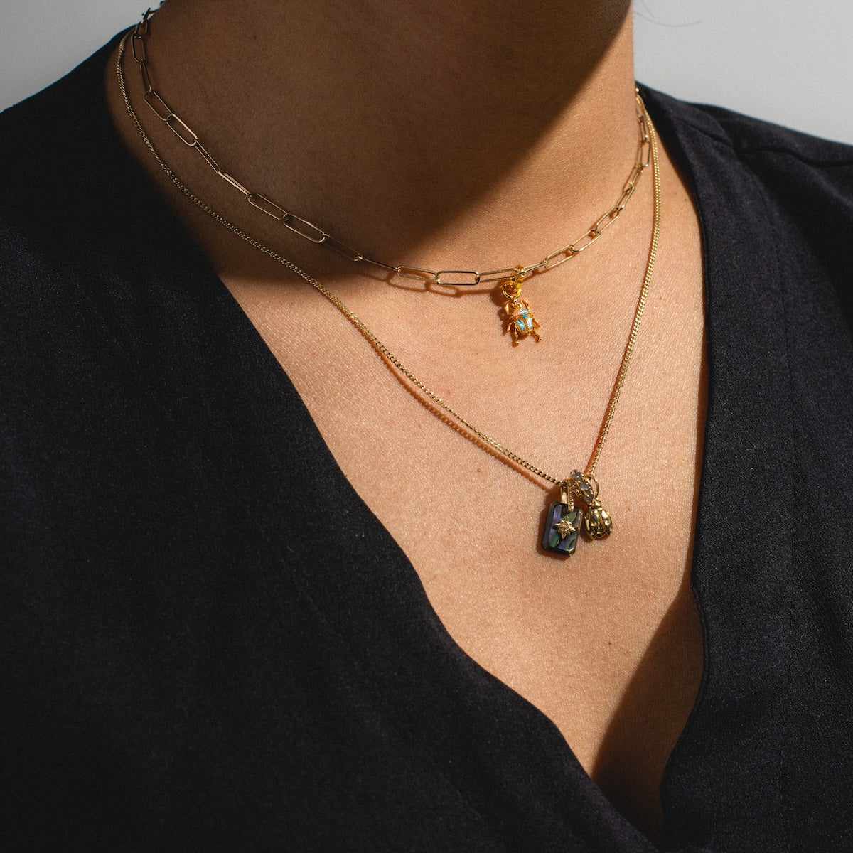 A close-up of a person wearing a black V-neck shirt and three gold necklaces from Charms by Girls Crew: an 18k yellow gold chunky chain with star charm, plus two finer chains with CZ stones and a Beetlejuice Charm.