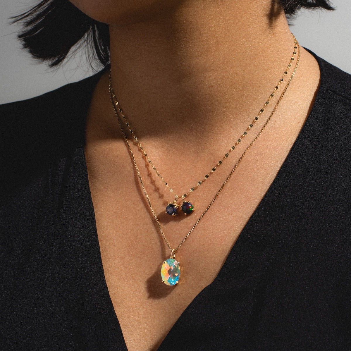 A person wears a black V-neck top and two layered gold necklaces—one with colorful beads, the other featuring the One of a Kind 14k Small Black Welo Opal Charm pendant.