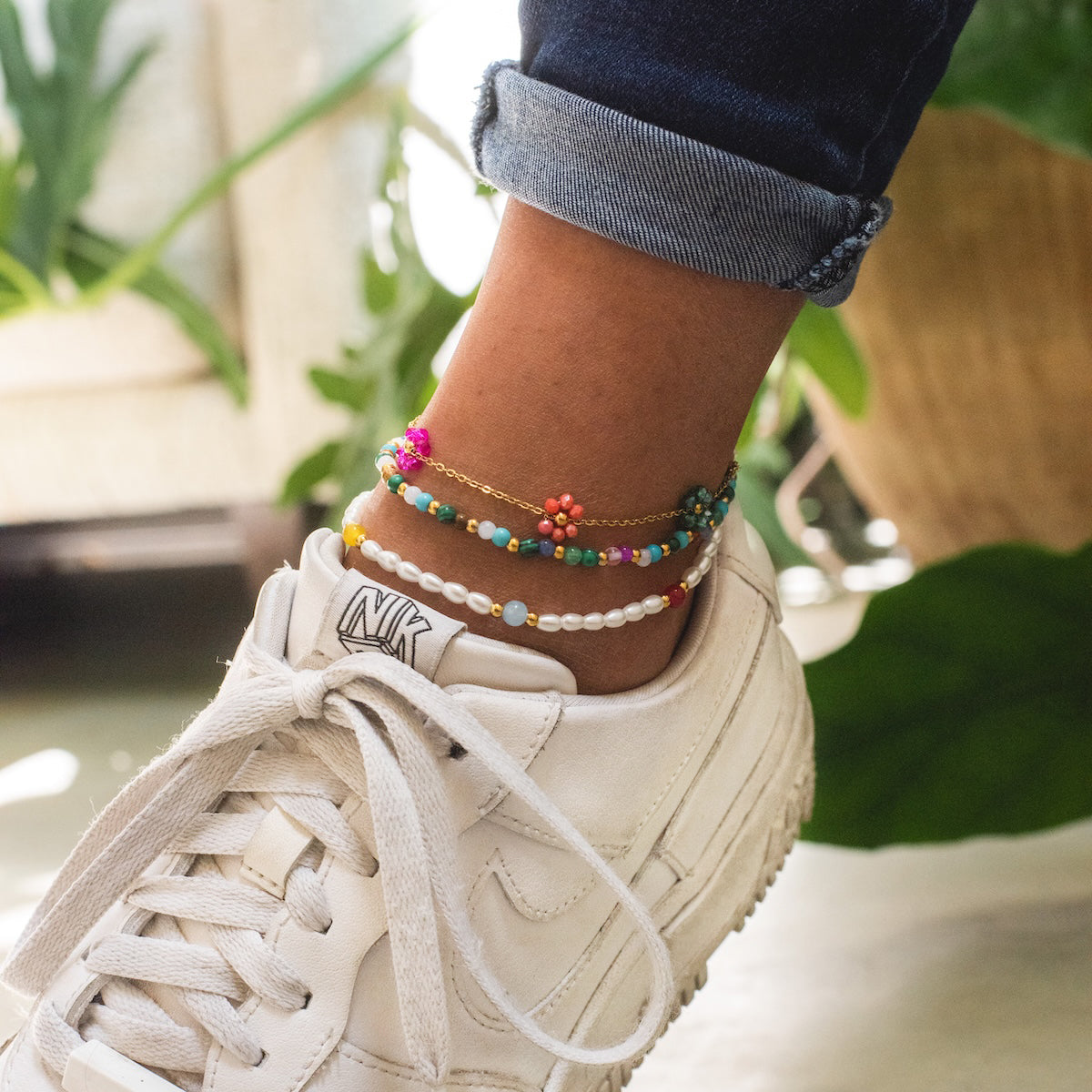 Close-up of a person wearing white sneakers, blue cuffed jeans, and the Pearl and Colorful Beaded Anklet—featuring pearls, bright beads, and floral accents—with green plants in the background.