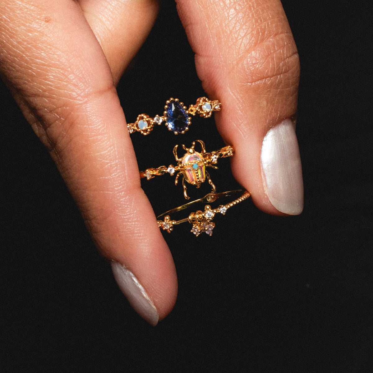 A hand with light polish holds three 18k yellow gold plated rings—a CZ stone band, the Beetlejuice Ring with a jeweled beetle design, and a small star ring—displayed against a black background.