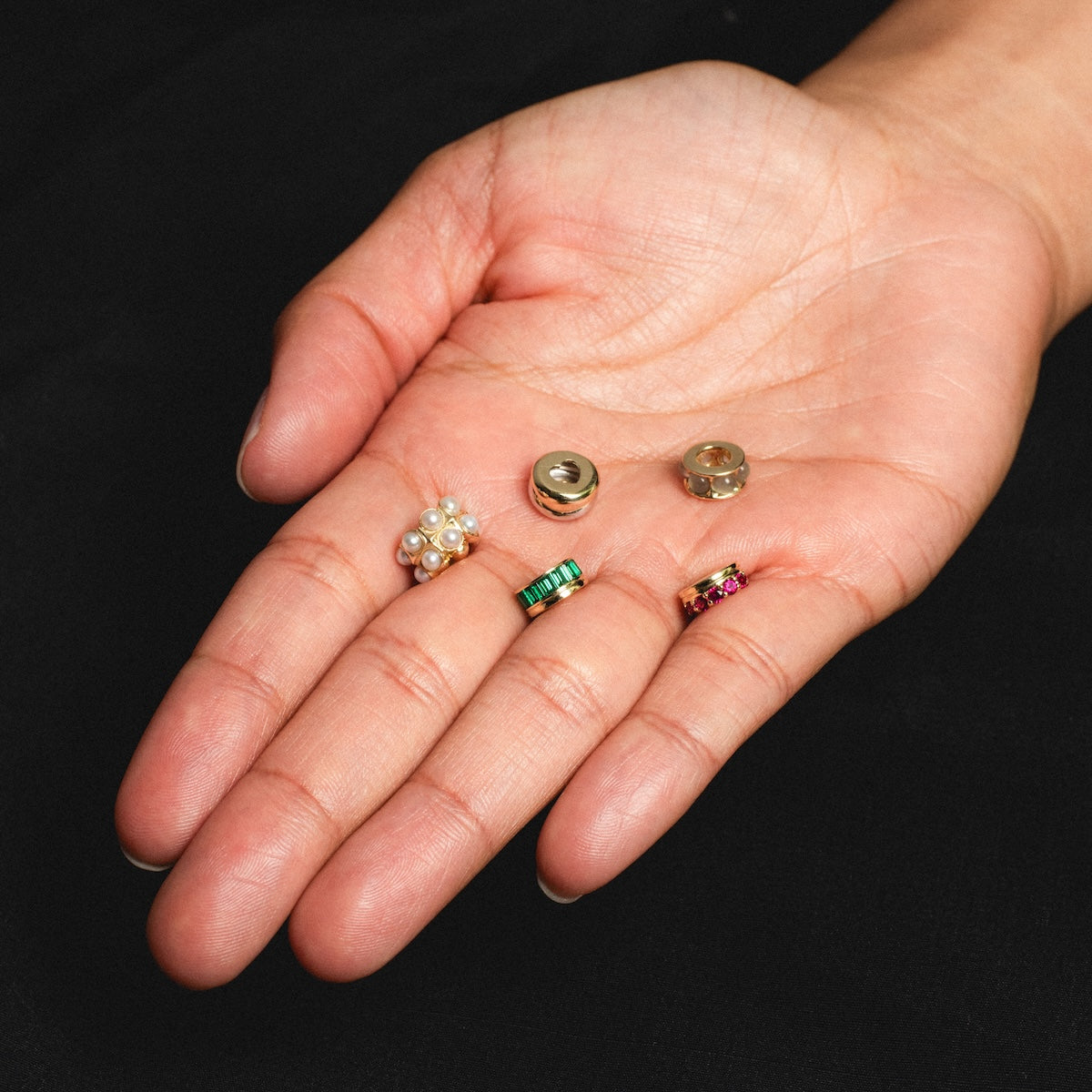 A hand with palm up holds five unique earring backs—including a pearl cluster, gold and clear discs, and gemstone bands—ideal for pairing with the Solid Gold Anniversaire Charm, set against a black background.