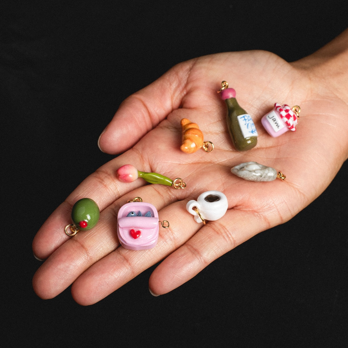 A hand showcases Charms by Warm Clay Co—hand-crafted croissant, wine bottle, baguette, olive, pink purse, coffee cup, jar, and tulip charms—against a black background.