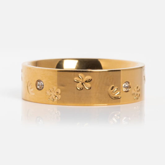The Work of Art Ring, a yellow gold plated piece with engraved flower and hand motifs and small round gemstones, is shown on a reflective white surface.