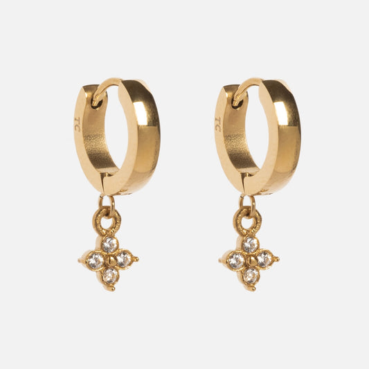The Merveilles Hoops are gold huggie earrings featuring dangling four-petal flower charms, each petal set with a clear gemstone. The floral bejeweled earrings are displayed against a plain white background.