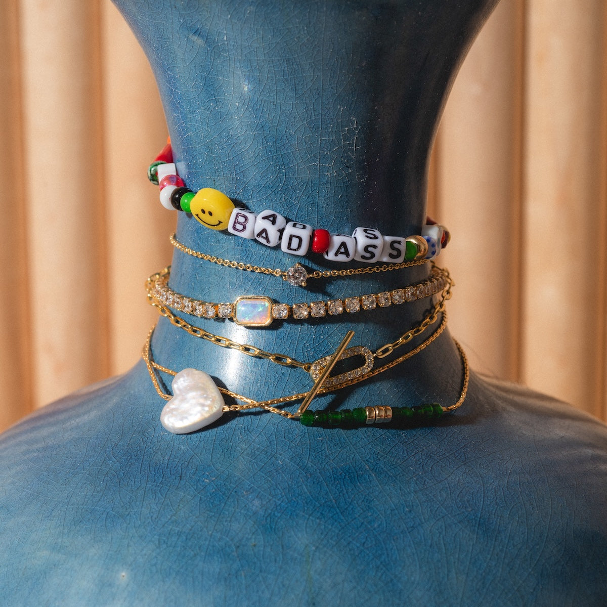 A blue vase displays five necklaces, including a handmade beaded “Bada$$” bracelet, a heart pendant, and gold plated chains with gems and green beads, against a beige striped background.
