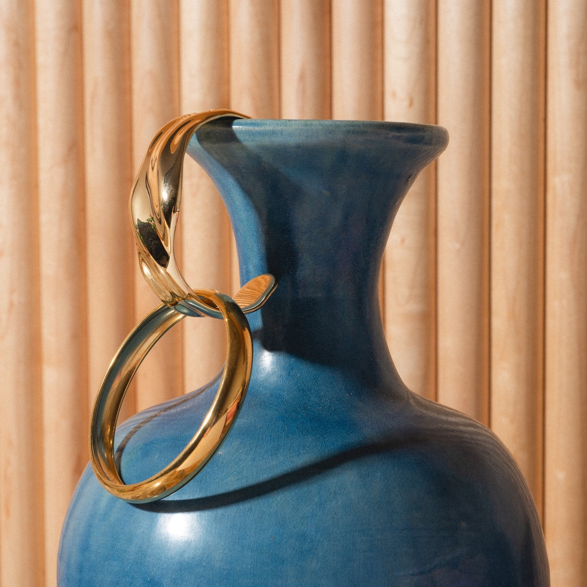 The Enza Small Bangle, crafted in shiny 14k gold, hangs from the rim of a blue ceramic vase against a backdrop of vertical light wooden slats.